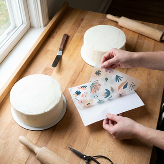 Hands peeling a printed edible icing sheet from its backing next to a frosted cake